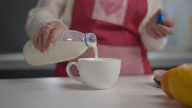 Close-up white cup on countertop with senior female hand pouring milk inside. Unrecognizable blurred Caucasian woman drinking dairy for breakfast in the morning at home indoors
