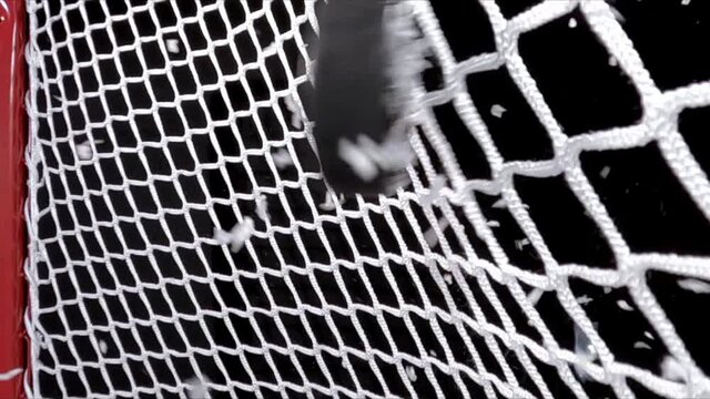 A close-up view from inside the goal net of an Ice Hockey puck hitting the back of it as shavings fly by, against a black background. Scoring a goal in ice hockey.