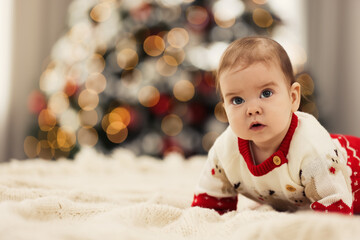Fototapeta na wymiar Cute little baby in Christmas sweater on knitted blanket against blurred festive lights, space for text. Winter holiday