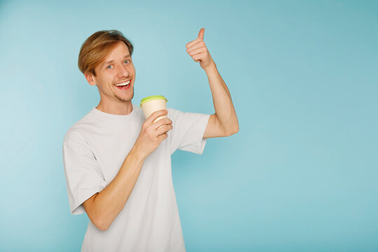 Young happy man standing over blue background smiling with happy face showing hand ok sign and drinking tea or coffee. Copy space for text.