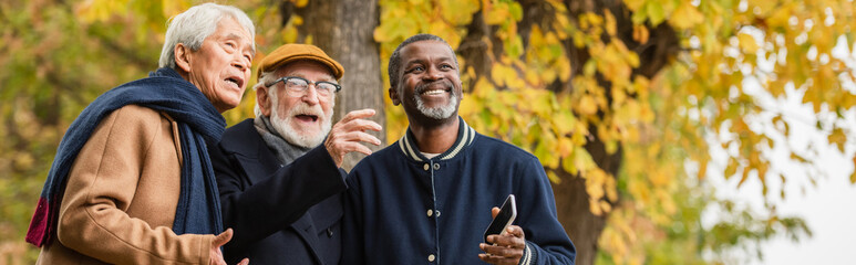 Smiling multiethnic elderly men with cellphone looking away in autumn park, banner.