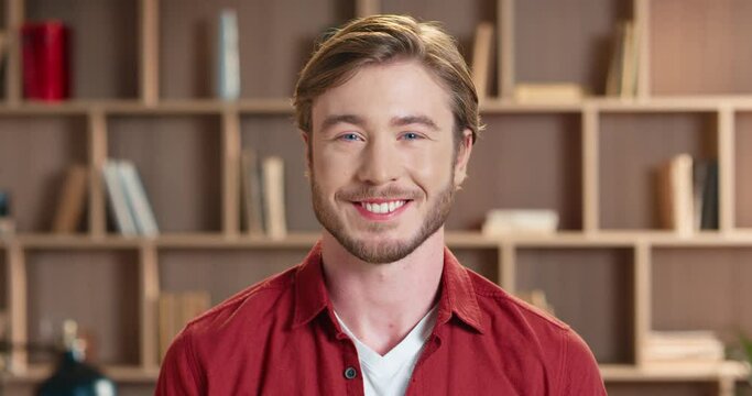 Close up portrait of handsome caucasian man with blue eyes in red shirt standing on bookcase background and smiling at camera.