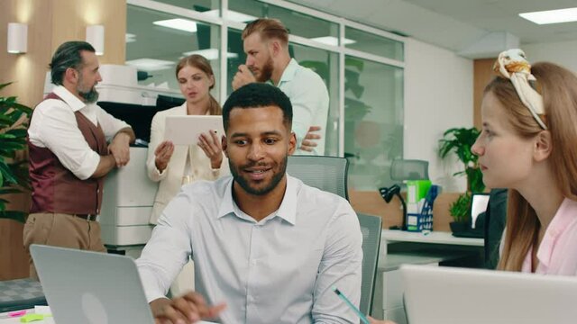 A blonde woman wearing a bandanna at work is talking to her handsome black worker, while in the back by the printer three colleagues are conversing amongst themselves. 4k