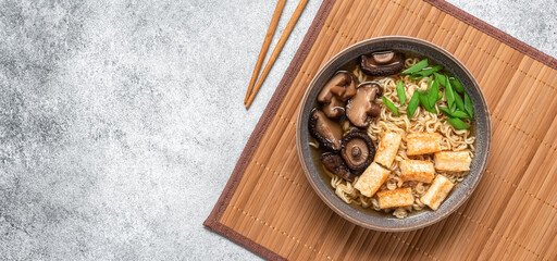 Asian vegan ramen soup with noodles, tofu cheese and shiitake mushrooms in a bowl on a bamboo mat. Gray concrete grunge background. Top view. Banner