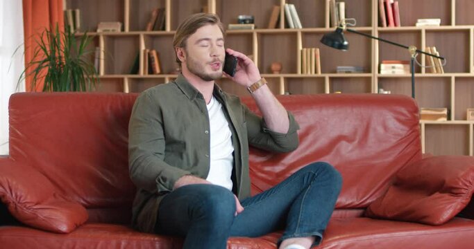 Successful Caucasian blond man freelancer sitting on stylish red sofa in modern office during short break and talking on cell phone.