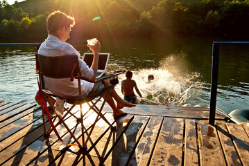 A man with a laptop and a glass of alcohol sitting in a chair. The children relax on the pier and jump into the water. The concept of summer, recreation, responsibility and supervision.