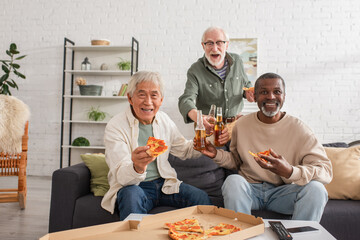 Happy multiethnic senior people holding pizza and beer bottles at home.