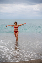 A middle-aged woman with a good figure in a red swimsuit on a pebble beach, running along the shore in the foam of the waves