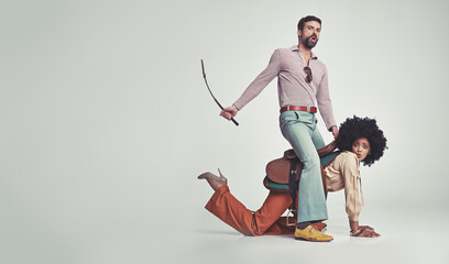 He's in charge now. A studio shot of an attractive man in 70s wear riding a young woman wearing a saddle while using a riding crop.