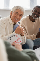 Smiling asian man holding playing cards near interracial friends at home.