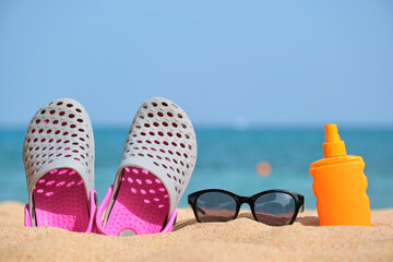 Fototapeta na wymiar Closeup of clogs shoes, sunscreen and black protective sunglasses on sandy beach at tropical seaside on warm sunny day. Summer vacation concept