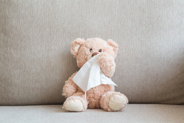 Brown teddy bear sitting on sofa and wiping nose with white paper napkin. Cold and flu virus. Children healthcare concept. Closeup. Front view. - obrazy, fototapety, plakaty