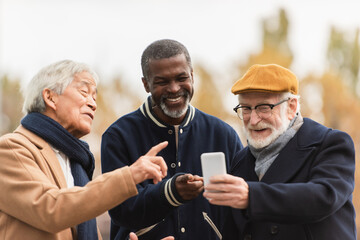 Positive interracial pensioners pointing at smartphone on urban street.