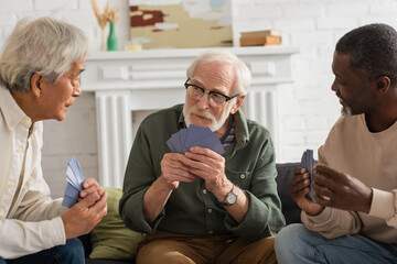 Elderly man holding playing cards near multicultural friends at home.