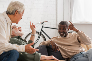 Cheerful multicultural elderly men talking on couch at home.