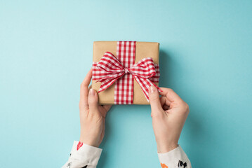 First person top view photo of valentine's day decorations young girl's hands in white shirt unpacking craft paper giftbox with checkered ribbon bow on isolated pastel blue background with empty space