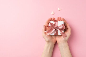 First person top view photo of valentine's day decorations female hands holding small white giftbox with pink ribbon bow on palms and two pink hearts on isolated pastel pink background with copyspace