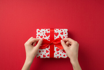 First person top view photo of st valentine's day decorations female hands tying red bow on giftbox in white wrapping paper with heart pattern on isolated red background with copyspace
