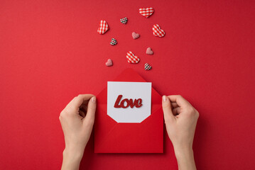 First person top view photo of valentine's day decorations girl's hands holding open red envelope with paper sheet inscription love and checkered hearts on isolated red background