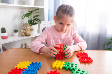 A little girl of 6-7 years old in a pink T-shirt sits at a table with a teddy bear at home, collects puzzles-constructor.