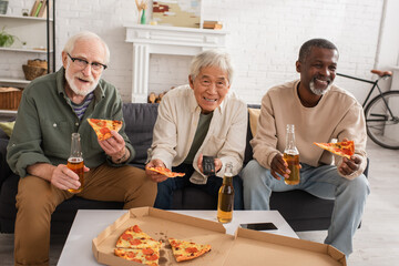 Positive interracial senior friends holding pizza and beer while watching tv at home.