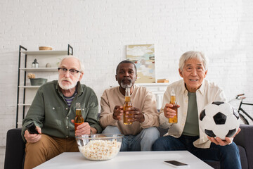 Senior multiethnic friends holding beer and football near popcorn at home.