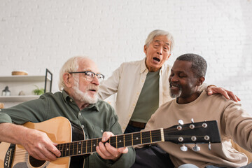 Multicultural pensioners playing acoustic guitar and singing in living room.