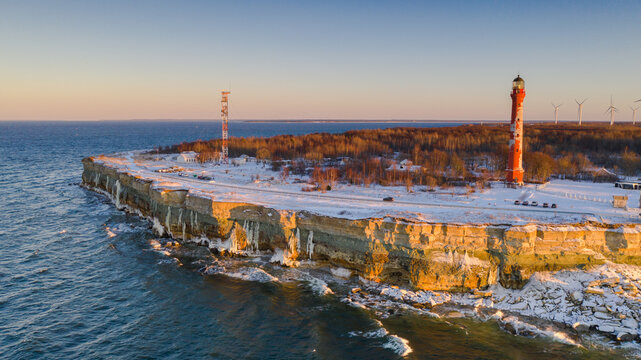 Aerial view to the winter sunset colored sea cape with lighthouse and wind turbines. The soft limestone cliff is leaking the groundwater and creating long icicles.