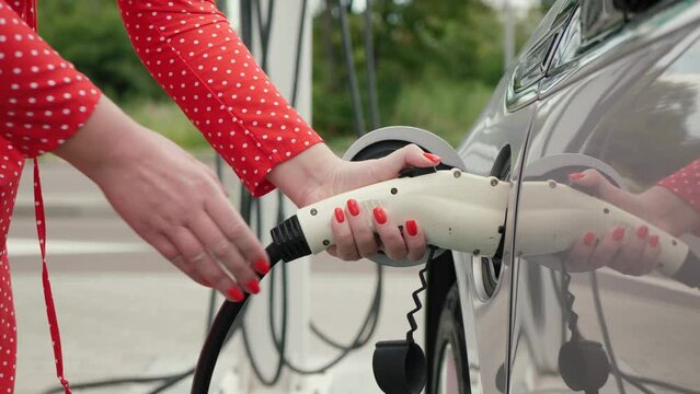 Unrecognizable woman in red dress with white dots approaches an electric car and connects plug. Lady plugging in power supply cable to charge EV, against charging point backdrop