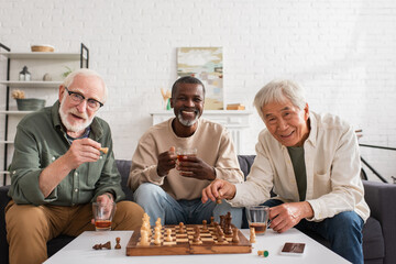 Cheerful interracial friends holding tea and chess figures at home.