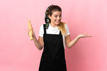 Young blonde woman holding a rolling pin isolated on pink background having doubts while raising hands