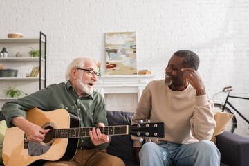 Senior man playing acoustic guitar near smiling african american friend on couch at home.