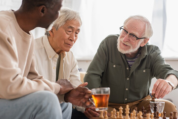 Smiling man playing chess with interracial friends near tea at home.