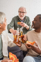 Smiling senior man clinking beer with interracial friends holding blurred pizza at home.