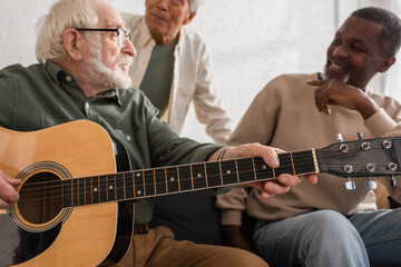 Acoustic guitar in hands of senior man playing near multiethnic friends at home.