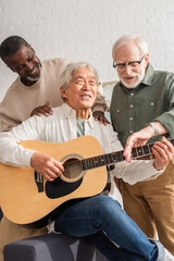 Happy senior multiethnic men standing near asian friend playing acoustic guitar at home.