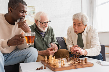 Interracial elderly friends holding tea while playing chess in living room.