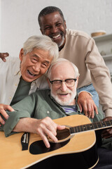 Cheerful interracial senior friends playing acoustic guitar and hugging at home.