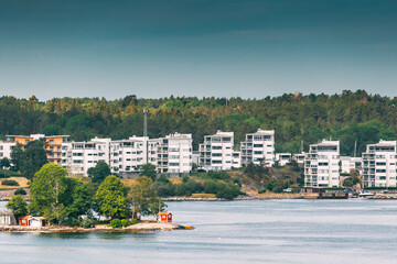 Sweden. Beautiful Red Swedish Wooden Log Cabin House On Rocky Island Coast In Summer Sunny Evening. Suburb With Multi-storey Houses On Background. Sweden. Beautiful Red Swedish Wooden Log Cabin House
