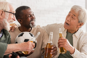 Excited interracial senior friends holding beer bottles and football at home.