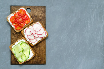 Assorted breakfast sandwiches with a slice of whole grain dark bread cream cheese cucumbers radishes cherry tomatoes on a gray concrete background on a cutting board. Flat lay. vegan diet post