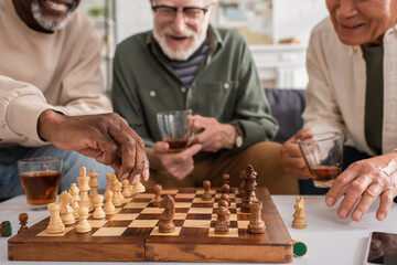 African american man playing chess near smartphone, tea and friends at home.