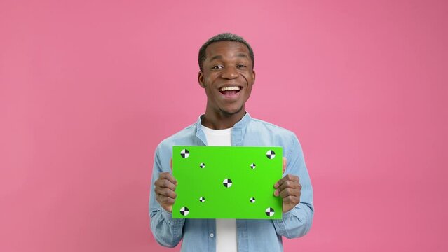 Happy African-American holding Banner with Green Screen Tracks Points for Copy Space. Empty Green Screen Board. Rejoices and Emotionally Shouts With Space for Text or Advertising on Pink Background.