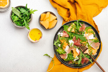 Summer melon salad with cantaloupe, prosciutto, soft cheese and arugula on white table background, top view, copy space