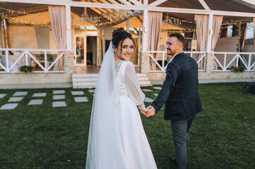 A young groom and a beautiful, smiling brunette bride in a white dress are walking along the green grass, in nature, against the backdrop of a cottage, at home. Wedding portrait, photography.