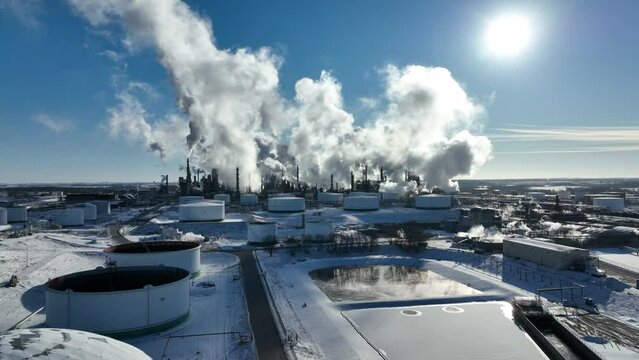 Petrochemical processing plant blocking out sun with smoke on otherwise clear winter day. Drone footage moving towards the refinery over tailing ponds and storage tanks.