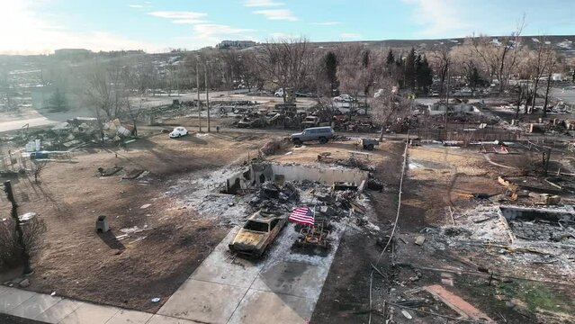 American flag flying amidst a neighborhood completely destroyed by a raging wildfire. Aerial 4K drone shot rising up and away to reveal the full devastation.