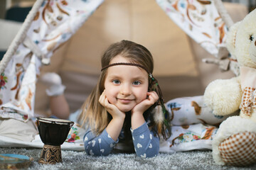 Portrait of charming little female kid leaning her head on hands while lying in wigwam and smiling on camera. Cozy home for playful time spending. © Sofiia Shunkina