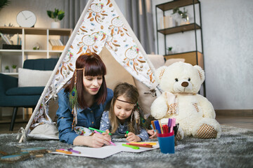 Happy caucasian mother and her cute daughter drawing near toy wigwam at home. Pretty woman and little child dressed in boho style. Playful time spending of family. © sofiko14