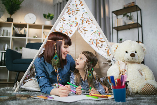 Smiling beautiful woman and cute little child lying together inside teepee tent and drawing with colorful markers in album. Mother and daughter spending free time at home with creativity.